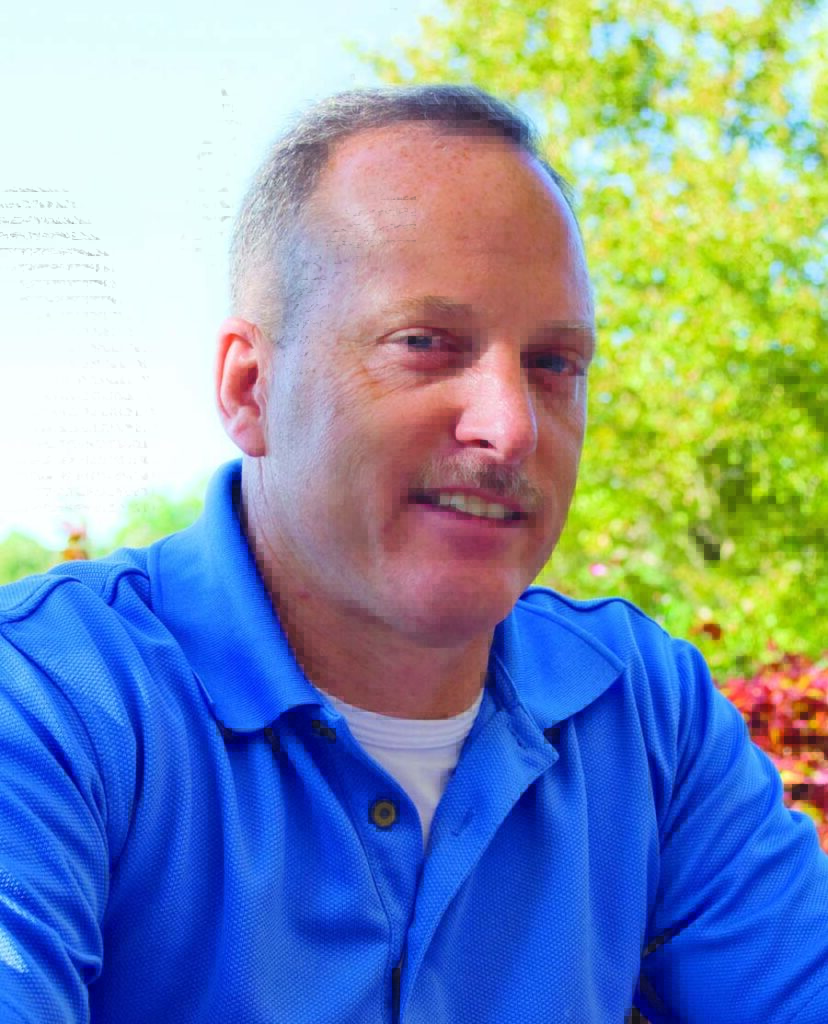 Portrait of James Donlan outside, wearing a blue polo with bright green trees behind him.