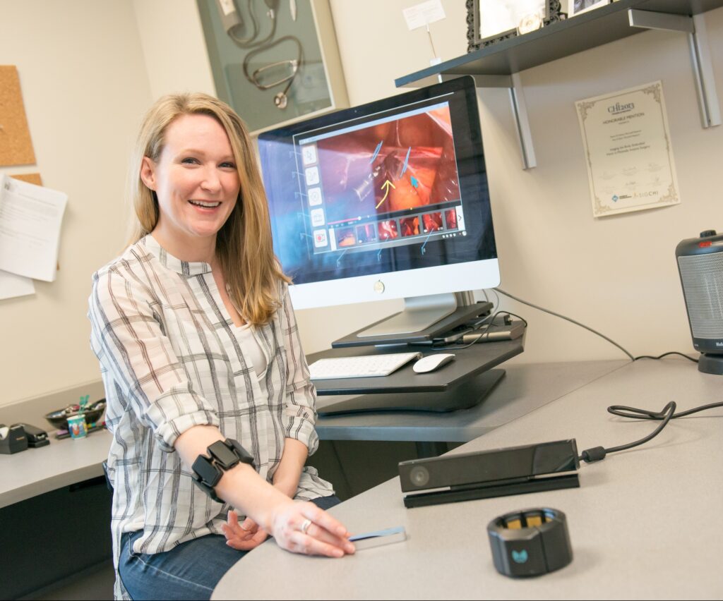 A woman with long blond hair wearing a white blouse with grey lines holds out her arm to show an electronic black bracelet . She is seated at a grey wrap around office desk with a flat screen monitor behind her.