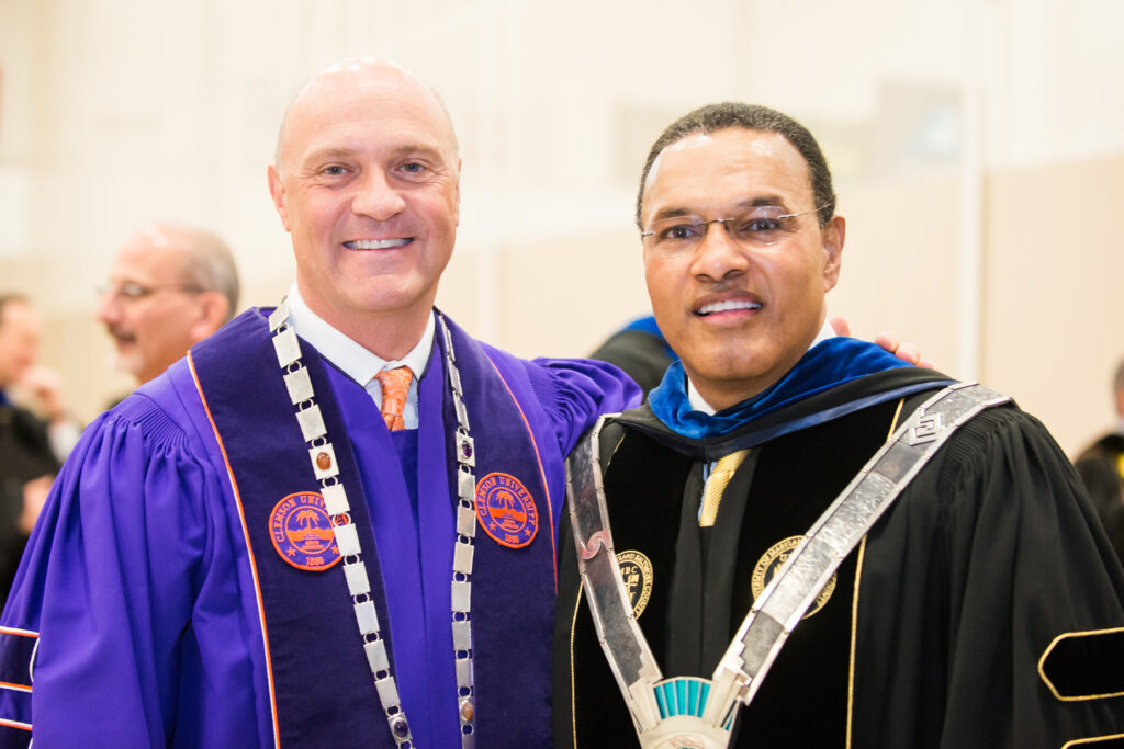 Two men stand in academic regalia, one in black and one in purple
