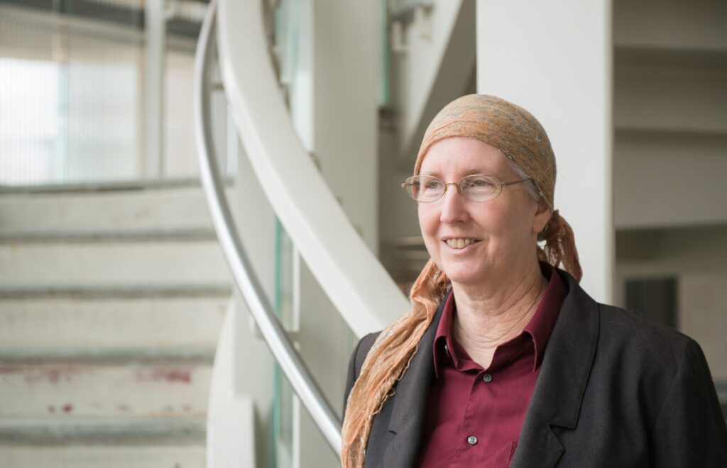 White woman with gray hair, a peach head scarf, and glasses stands in front of a staircase, smiling. She wears a burgundy button-up shirt and blazer.