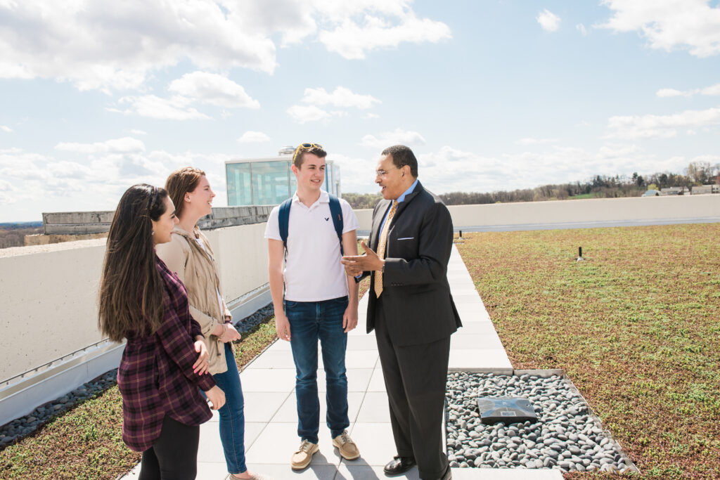 Man in suit stands with three students in casual clothes on a green roof with blue sky above.