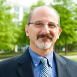 A man with a beard and mustache wearing dark rimmed glasses and a blue suit and striped tie smiles at the camera, there are green trees in the backgound