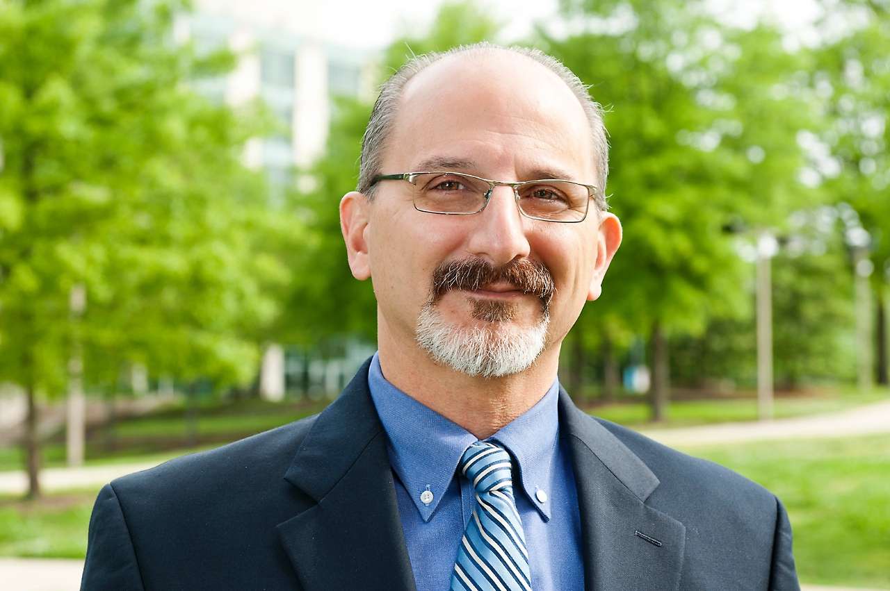 A man with a beard and mustache wearing dark rimmed glasses and a blue suit and striped tie smiles at the camera, there are green trees in the backgound