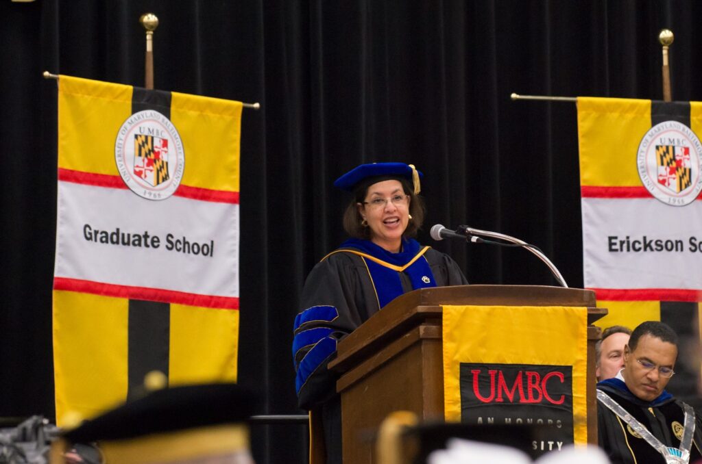 Middle-aged black woman in Ph.D. regalia delivers remarks at a podium. Podium reads 