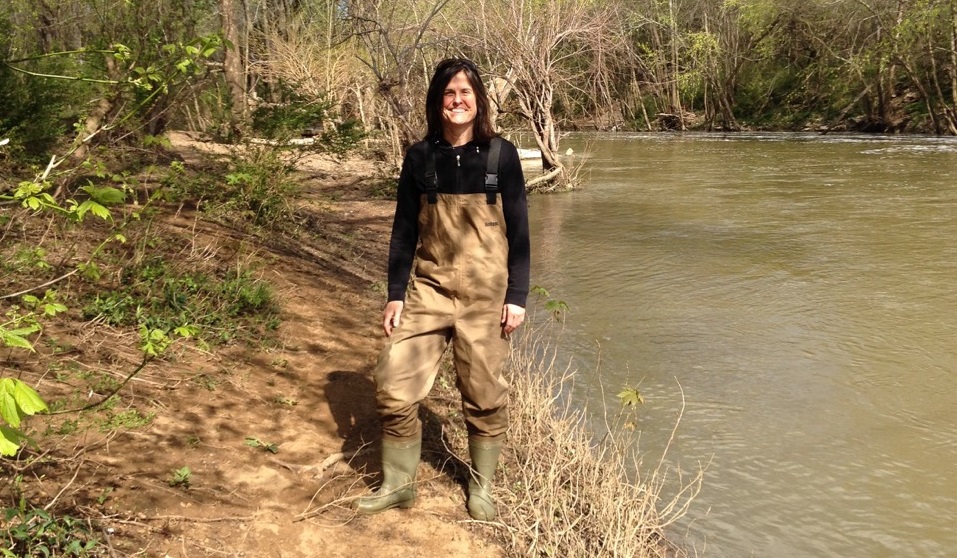 Middle-aged white woman with dark hair stands near water, wearing boots and waterproof overalls.