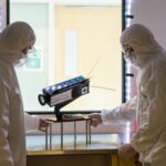 Two scientists in protective suits stand next to a piece of equipment with a large lens