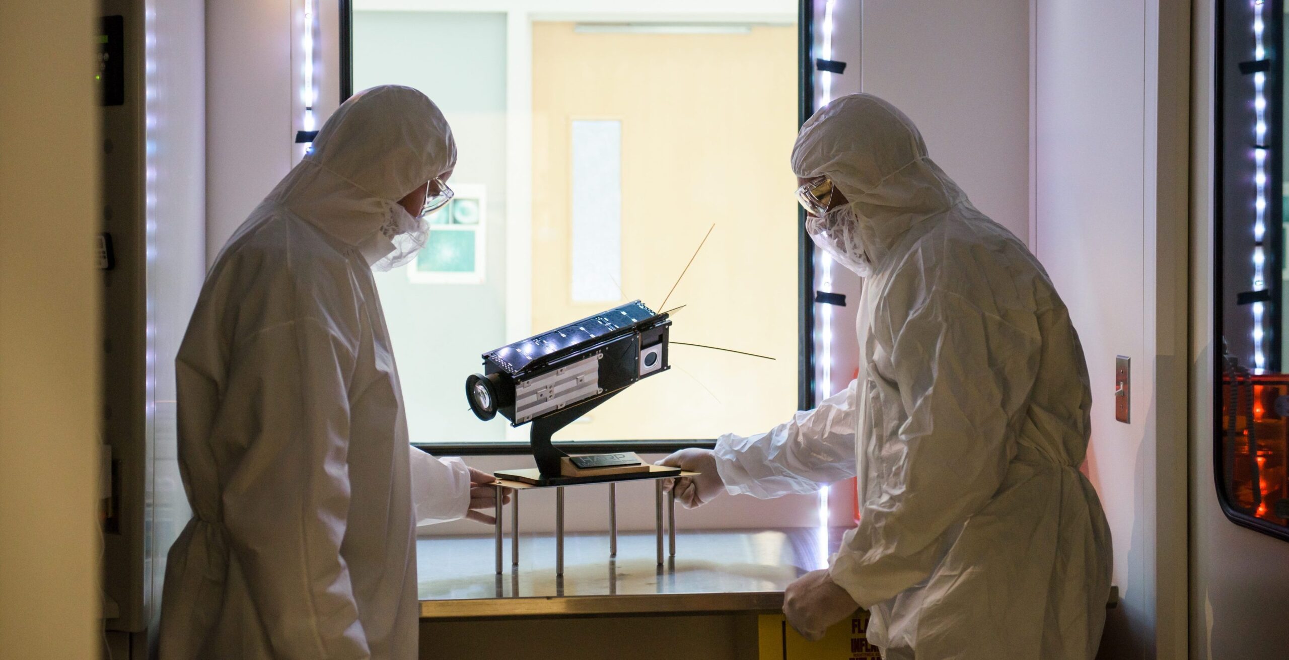 Two scientists in protective suits stand next to a piece of equipment with a large lens