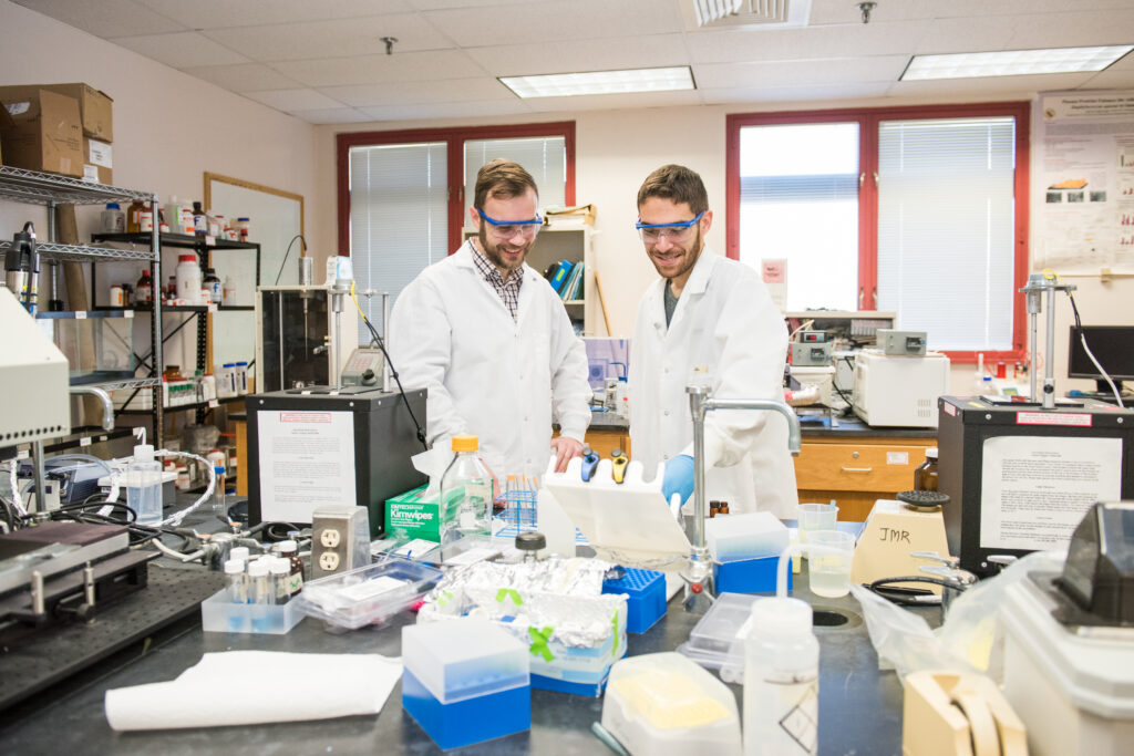 A profess and student do an experiment in a lab, wearing protective gear