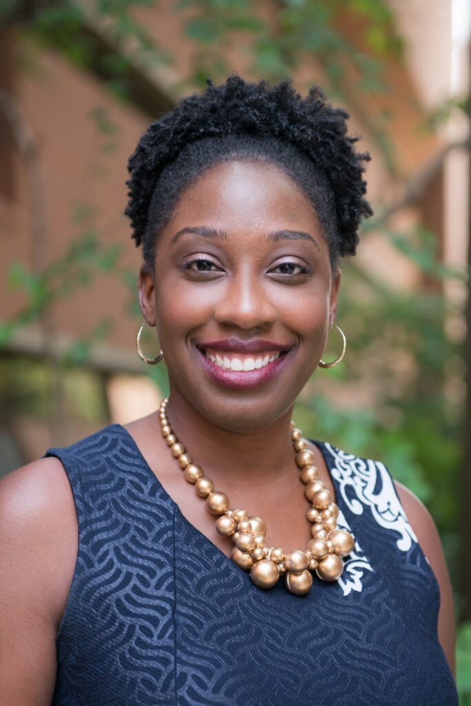 Outdoor portrait of smiling black woman wearing navy blue dress and gold necklace.