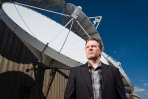 A portrait of David Tobin standing infront of a large satellite.