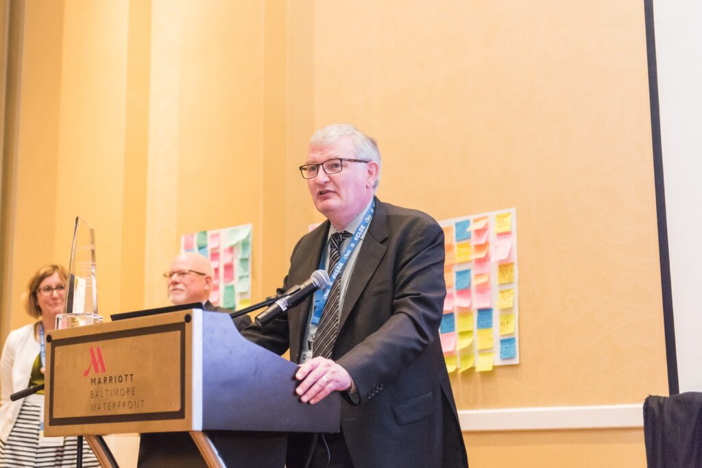 Man in suit stands at podium that holds a glass trophy. Multicolored post-its stick to the wall behind him.