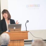 Women presents a talk from a podium, with laptop in front of her and projector screen behind her.