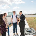 Man in suit stands with three students in casual clothes on a green roof with blue sky above.