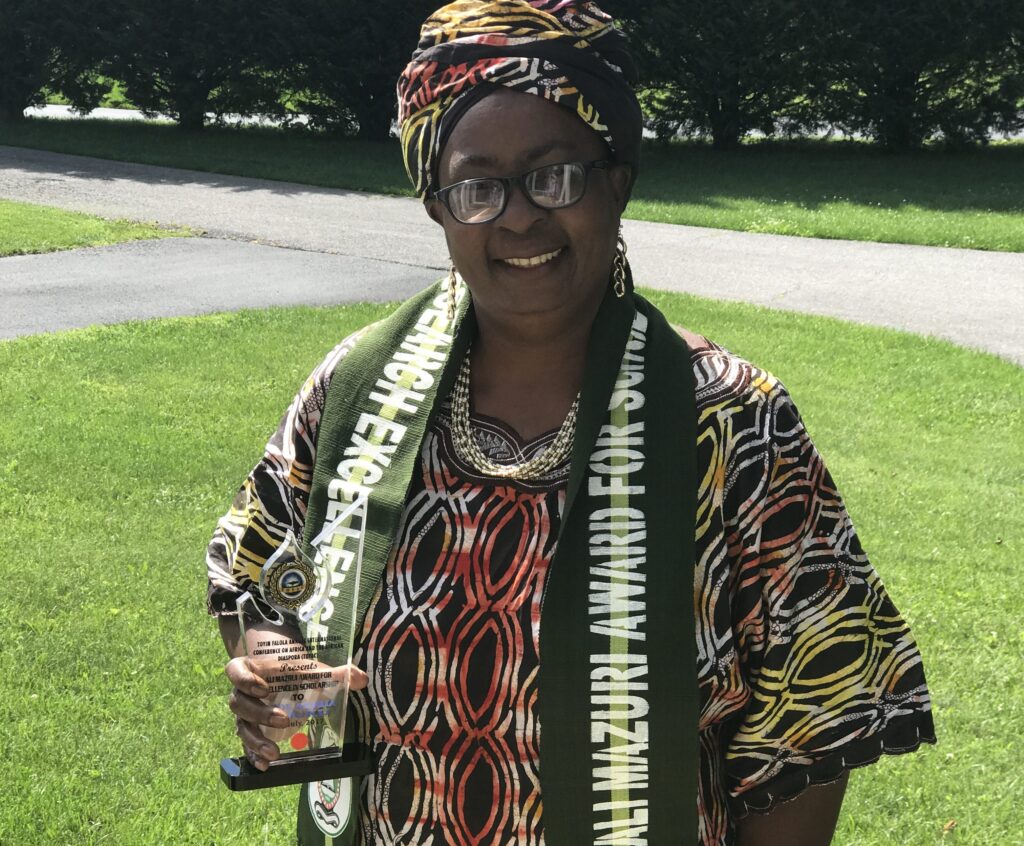 Adult woman wearing Nigerian clothing and head piece holds a glass award in her right hand as she smiles at camera, green fields and trees are behind her