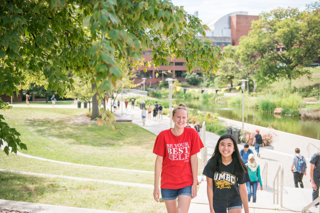 Two students walk near the library pond, one in a red shirt and one in a black shirt with UMBC athletics logo.