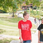 Two students walk near the library pond, one in a red shirt and one in a black shirt with UMBC athletics logo.