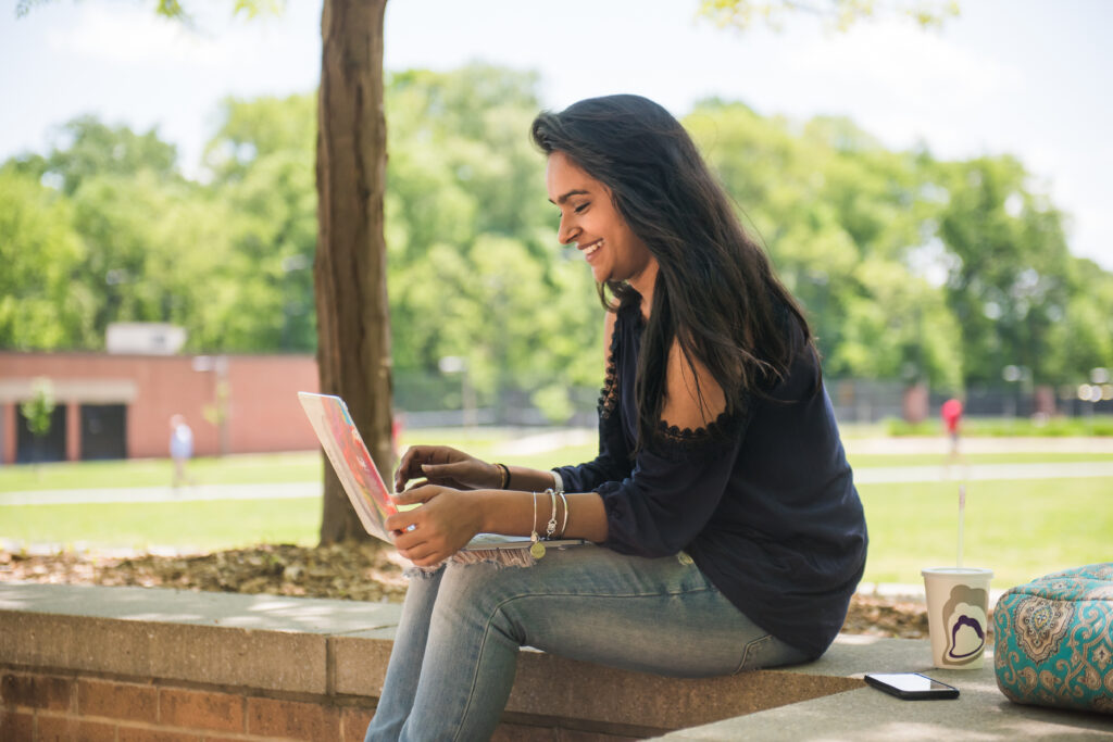 A student in jeans and a black shirt sits on a step working on a laptop, surrounded by grass and trees.