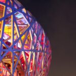 Photo of Beijing Olympic site, featuring metalwork in the shape of a web, with pink, orange, and yellow lights.