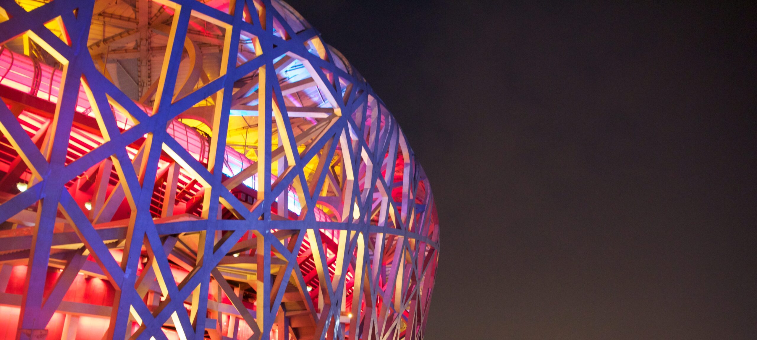Photo of Beijing Olympic site, featuring metalwork in the shape of a web, with pink, orange, and yellow lights.