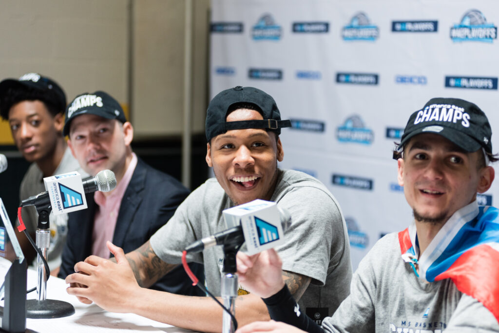 Basketball players in gray shirts sit behind microphones.