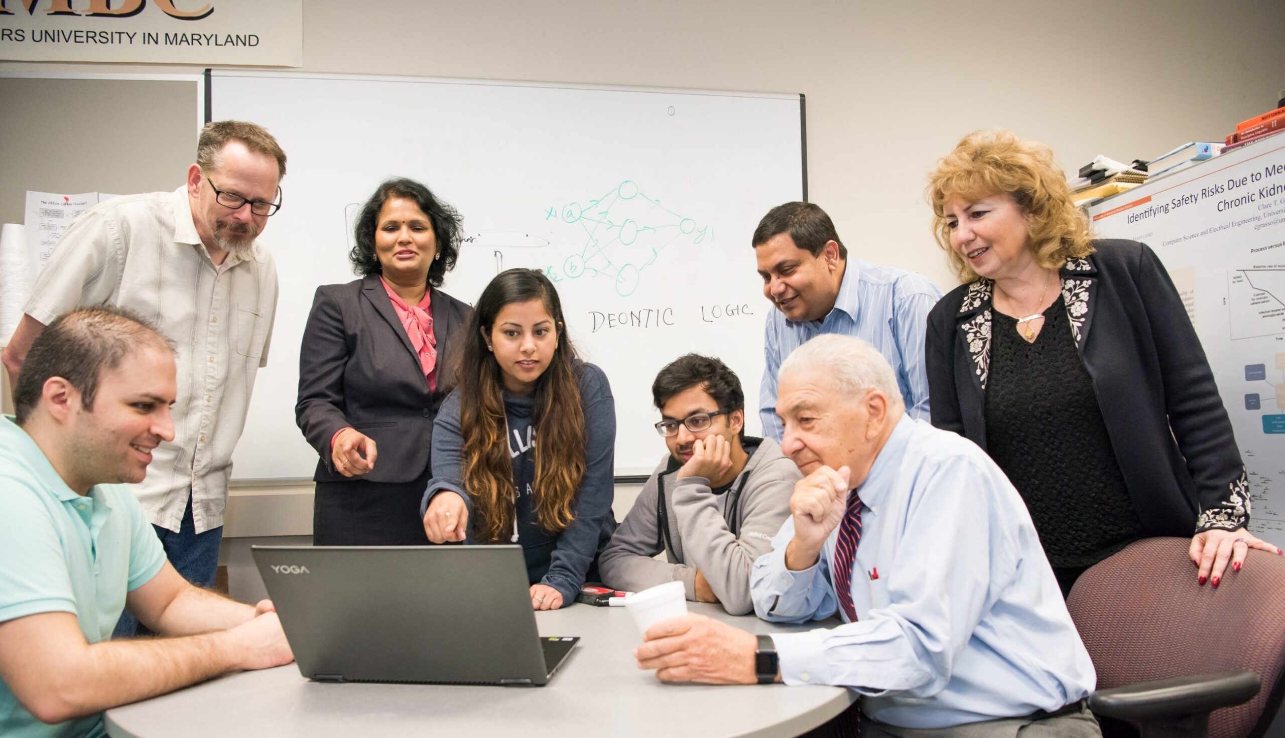 A diverse group of students and professors clusters around a laptop screen in a conference room.