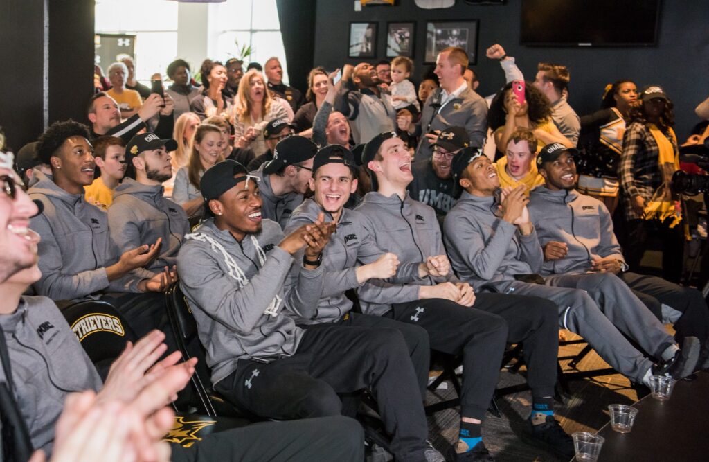 Basketball players seated, wearing gray jackets, smile while watching TV livestream.