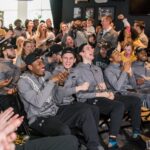 Basketball players seated, wearing gray jackets, smile while watching TV livestream.