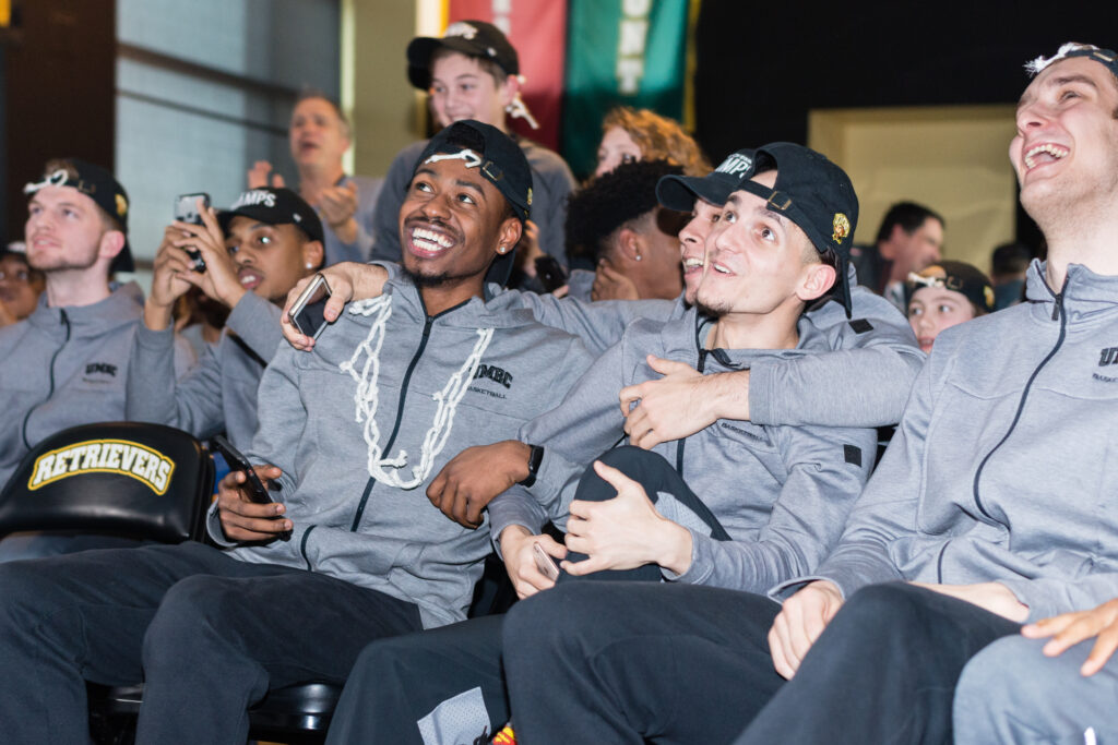 Basketball players in matching gray jackets and black hats celebrate, hugging each other.
