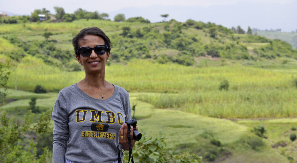 Women in sunglasses and UMBC Retrievers sweatshirt stands on a grassy hill.