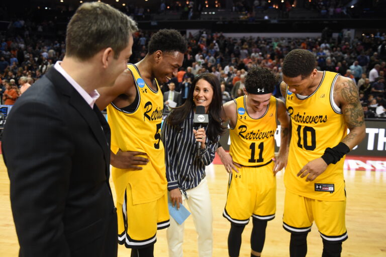 Journalist (center) holds microphone and speaks with coach and three players in yellow jerseys.