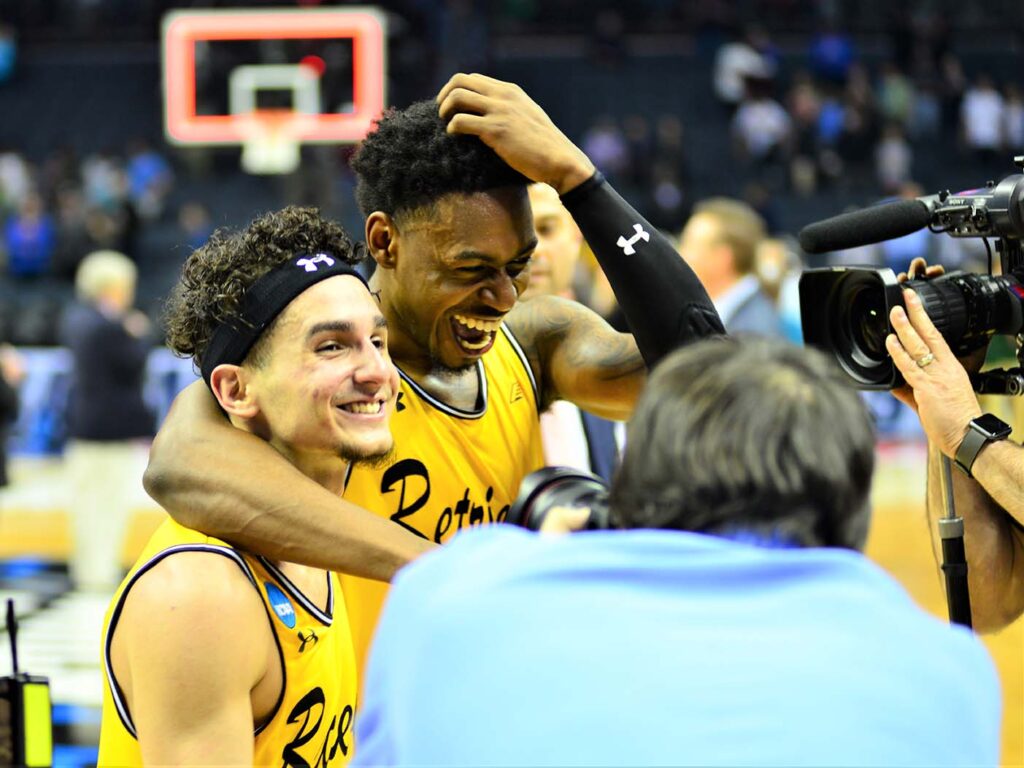 Two basketball players in gold jerseys smile and embrace.