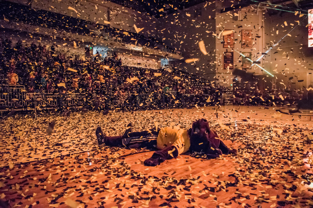 Person in retriever mascot costume and black and gold uniform celebrates by laying on a gymnasium floor, covered in confetti.