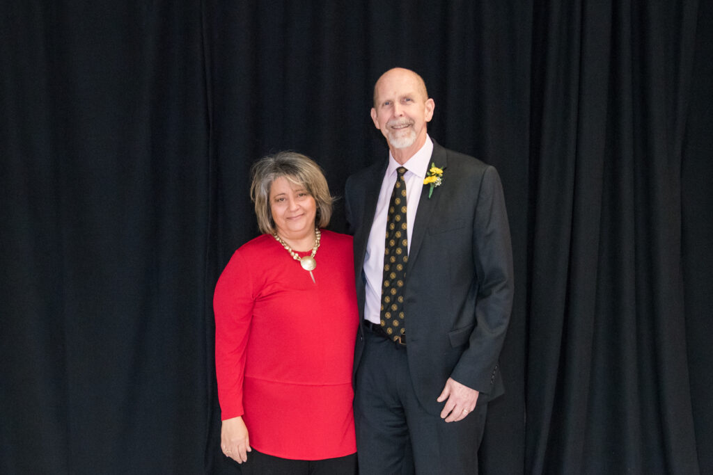 Woman in red shirt and man in suit with black and gold tie pose together.