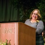 Woman in gray and black patterned dress stands at podium, smiling.