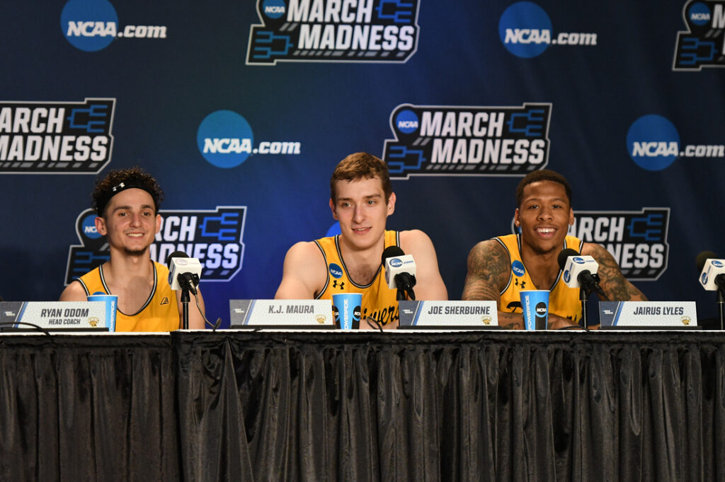 Three basketball players speak into microphones in front of March Madness backdrop.