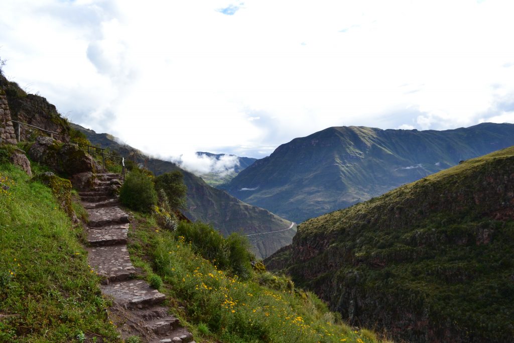 A vast landscape featuring mountains covered in greenery, a stone walking path, and a sky full of clouds.