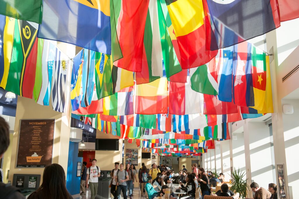 Many flags from countries around the world hanging from the ceiling in UMBC's Commons building. Students are walking and sitting at various tables.