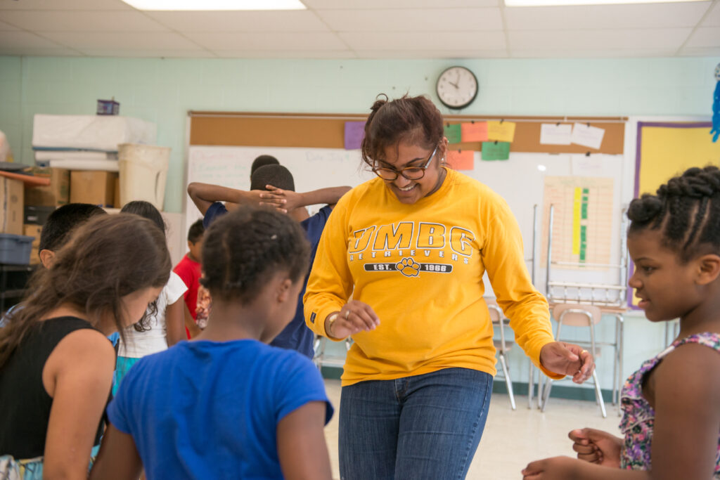 College student in yellow UMBC shirt leads elementary students in a movement-based activity.
