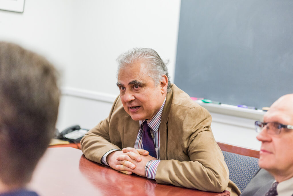 Man in a brown suit jacket and purple tie speaks at a conference table. Another man, in glasses, appears at right.