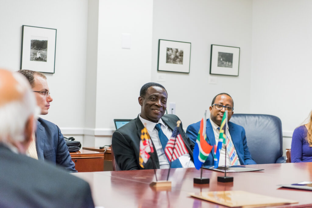Three men in suits sit at a conference table. Flags of Maryland, U.S., and South Africa appear on the table.