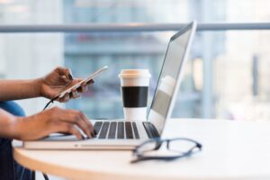 A laptop, a coffee cup, and glasses sit on top of a round wooden table. The hands of a person are hovering over the keyboard. One of the hands is holding a cell phone.