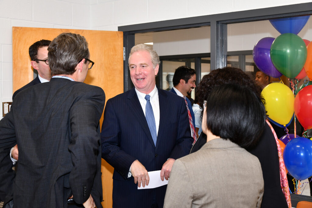 Man in suit at center of photo converses with a group. Balloons on the edge of frame.