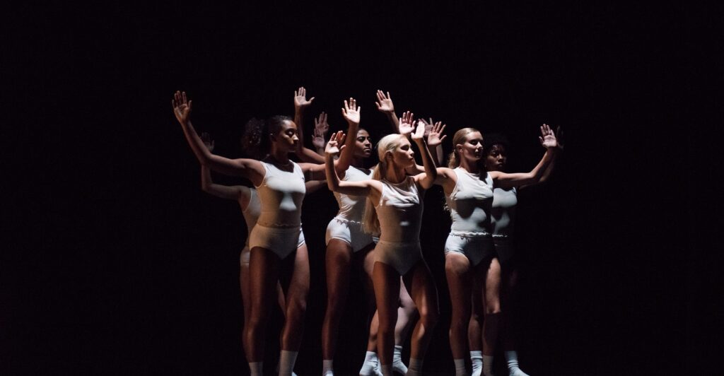 A group of women dancers performs, clustered together with arms in the air, with simple, white costumes and black background.