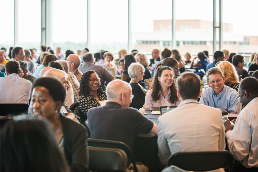 Hundreds of professionals sit around tables in a room, talking, in groups of 8-10