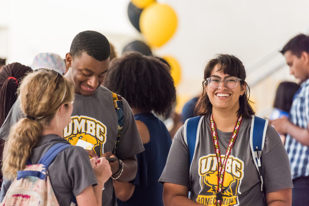 Three students wearing UMBC shirts cluster in a group, with one looking up toward the camera.