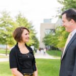 A man and woman in professional clothing stand outdoors in front of a manicured lawn and trees