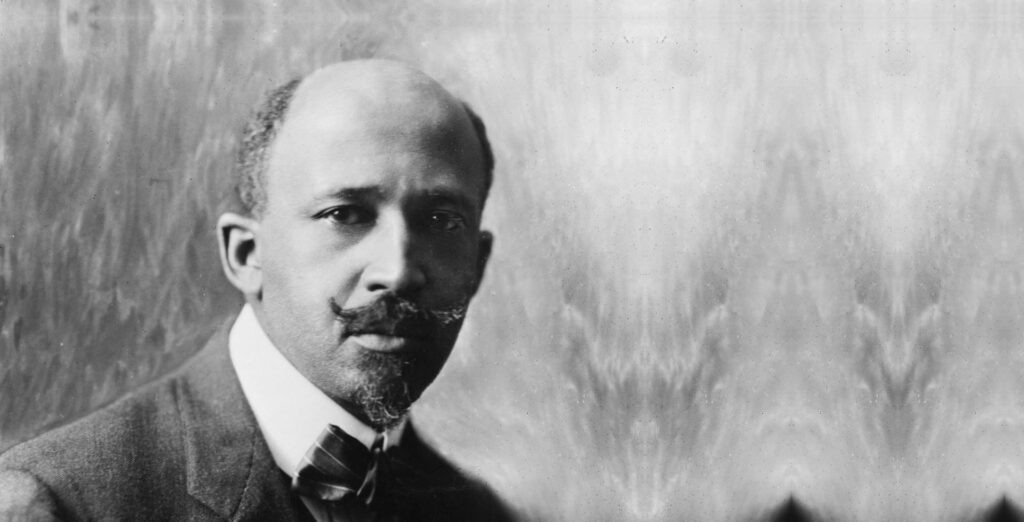 Historic black and white photo of a man with balding hair and handle bar mustache wearing a suit looks at camera.