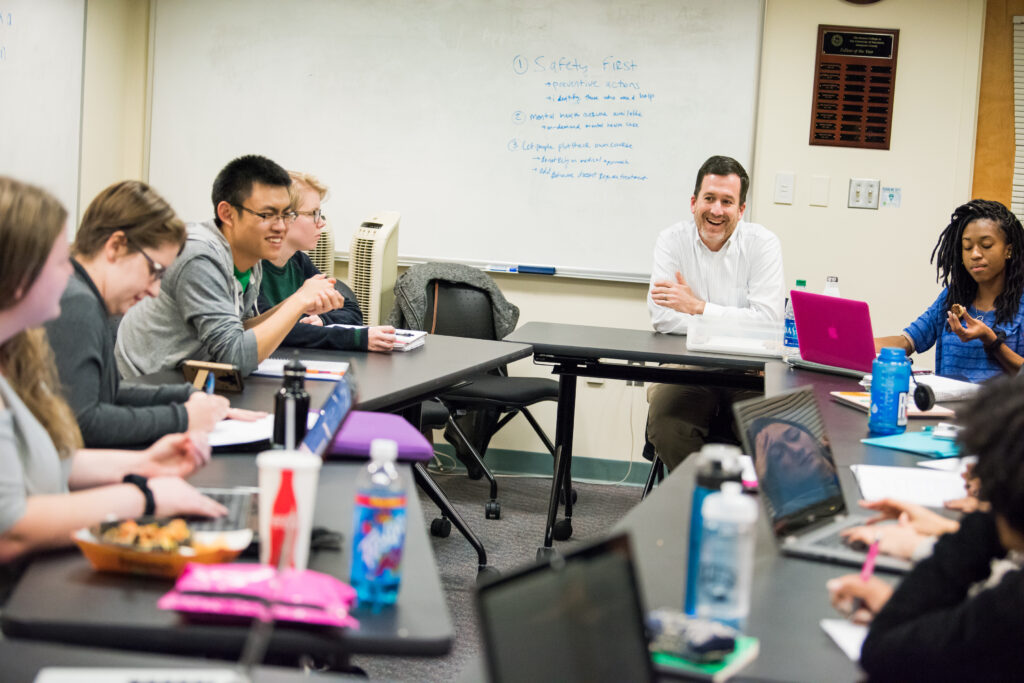 Teacher and a several students sit around tables set in the shape of a circle, eating snacks, typing on laptops, talking and smiling.