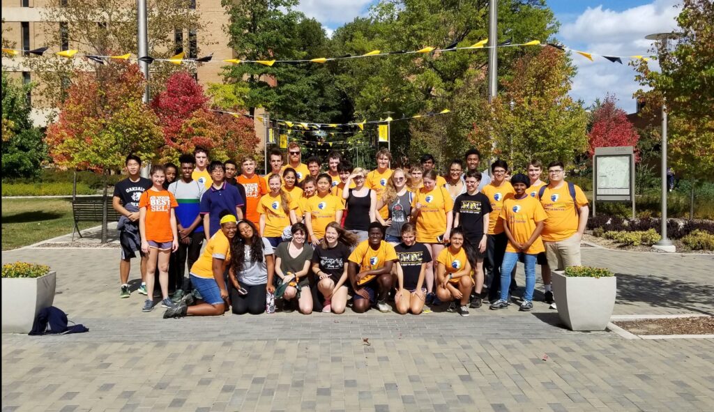 A group of UMBC students in gold t-shirts poses in front of campus.