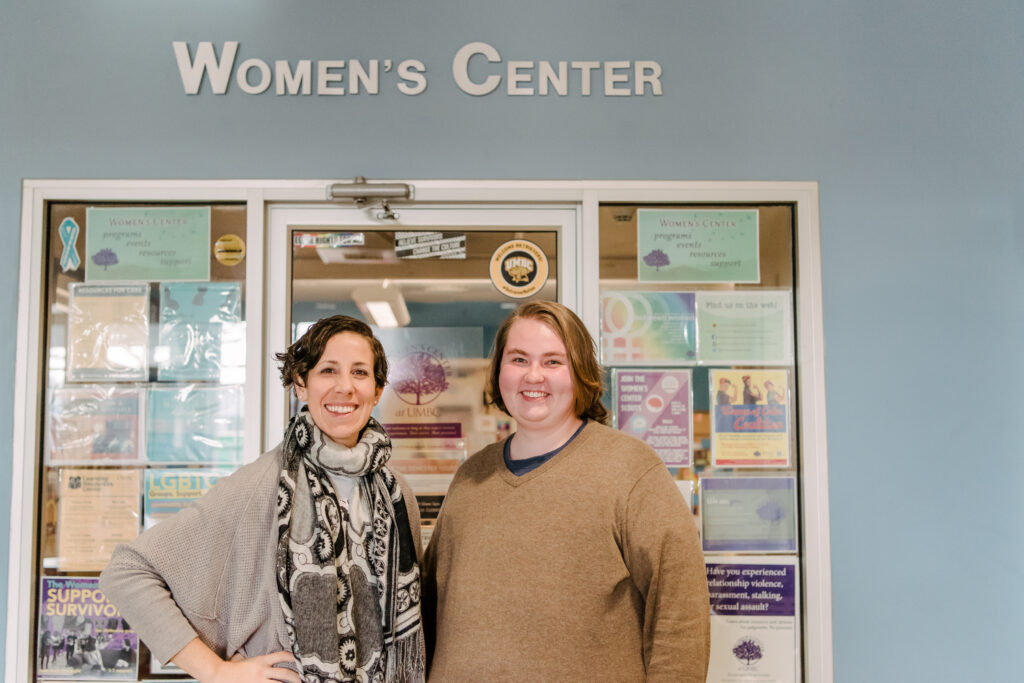 Two people -- one in gray sweater and patterned scarf and one in light brown sweater -- pose in front of the UMBC Womens Center.